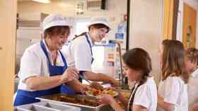 Two dinner ladies serve lunch to students queueing in a school canteen.