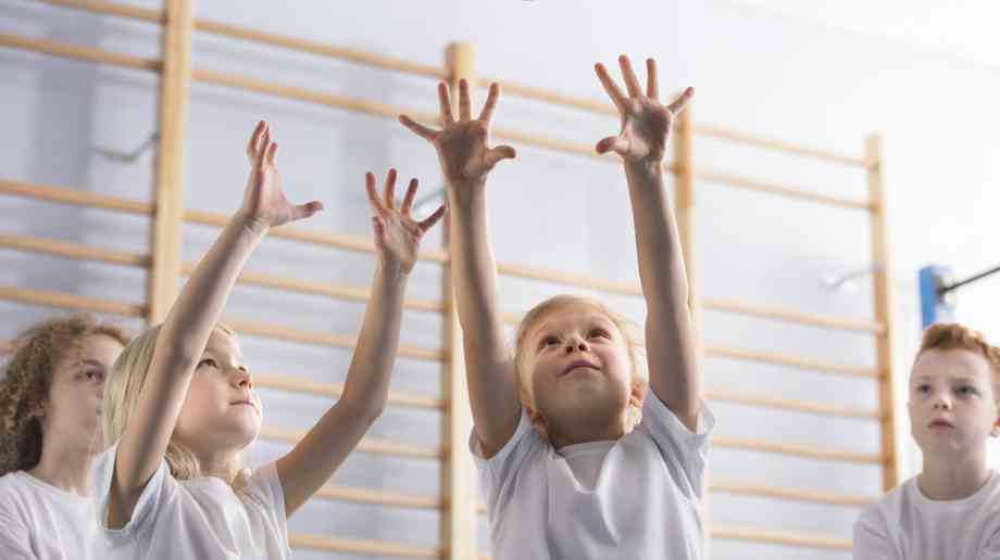 A group of children in white tee shirts play volleyball. A young girl is in the centre of the image with her arms outstretched to catch the blue and yellow ball. 