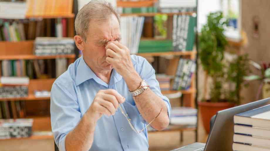 A man strains his face with stress. He is sat at a laptop.