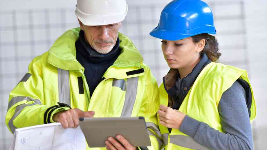 Female construction apprentice uses tablet next to older employee.