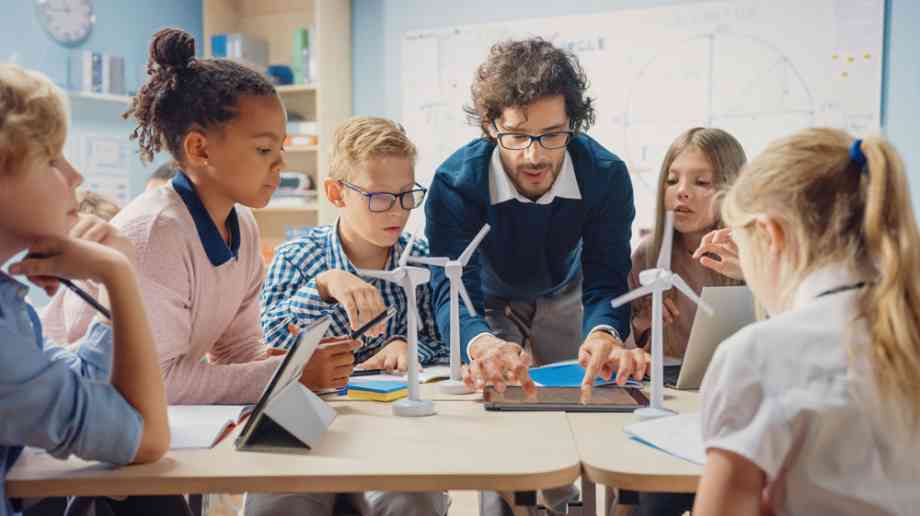 Teacher uses a tablet to teach four children around a table with model wind turbines.